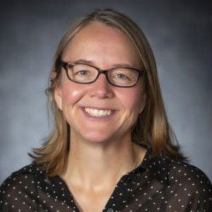 Ruth Sofield is smiling. She is light-skinned, has blond hair, and wears glasses and a brown shirt with beige polka dots.
