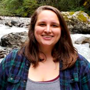 Sarah Brown is a light-skinned female with long brown hair and a warm smile. She is outside, by a river, and wears a plaid shirt over a grey T-shirt.