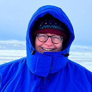 Pamela Miller is a light-skinned female who smiles and wears glasses, a knit cap, and a blue winter parka.
