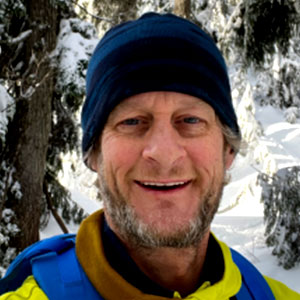 David Rossiter is a light-skinned, bearded, smiling male standing in a snow-covered forest and wearing a knit cap and bright green winter coat.