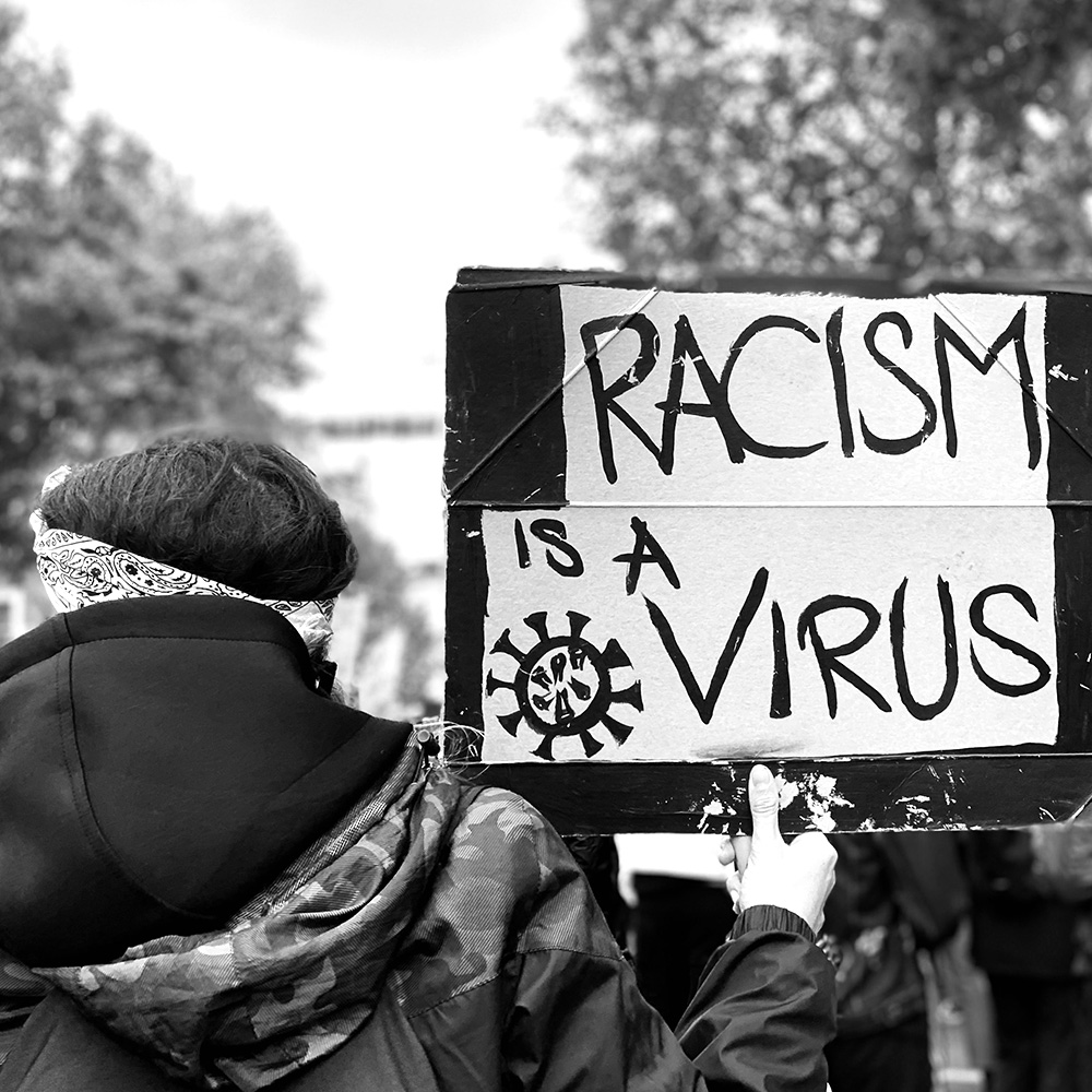 Protest with a protester holding up a sign saying RACISM IS A VIRUS.