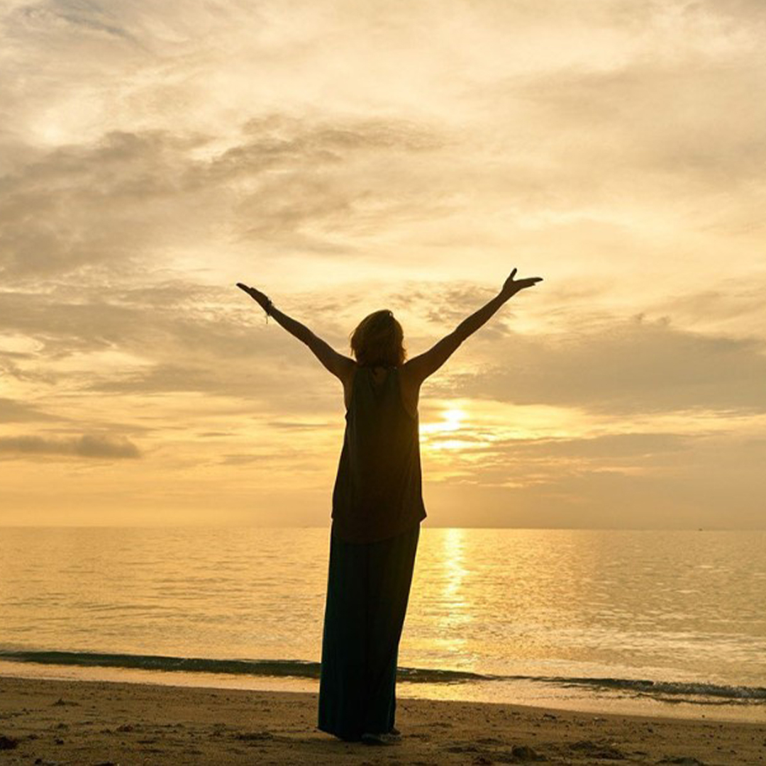 Person standing on the beach during sunrise with their arms raised