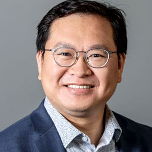 Speaker Maxwell Leung wears wire-rimmed glasses and has white skin, a broad, warm smile and short auburn hair. He is casually dressed in a light grey shirt and a slate blue linen blazer.