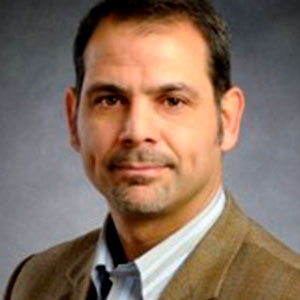 Vincenzo Bianco has brown eyes, short brown hair, white skin, short facial hair, and a serious expression. He wears a brown blazer and grey shirt.