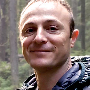 Close up of Daniel Donato's face. He has white skin, brown eyes, and a warm smile. Forest trees are in the background.