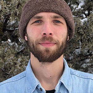 Chris Mulverhill has white skin, rosy cheeks, a brown beard and wears a knitted cap and light blue denim shirt.