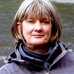 Andrea Geiger has white skin, a warm smile, short grey and blond hair with bangs, and wears an outdoor jacket and turtleneck. A river can be seen behind her.
