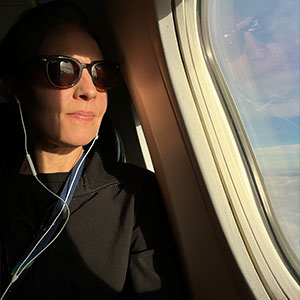 Ingrid Barrentine looks out of an airplane window. She has white skin and is wearing sunglasses, wired ear buds, and a black shirt.