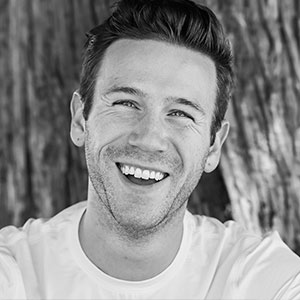 In this black and white portrait Jordan Stead smiles broadly and slightly tilts his head. He has white skin, short hair, short facial hair, and wears a T-shirt.