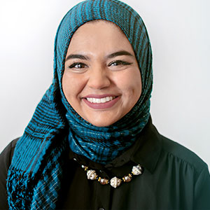 Sana Ullah smiles warmly. She is wearing a green head scarf and a necklace of large beads.