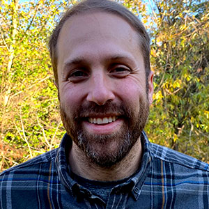 Gabe Epperson smiles warmly. He has white skin, brown hair and beard. He wears a blue flannel shirt. Vibrant green shrubs and trees are behind him.