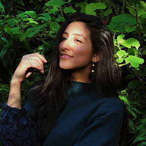 Diandra Marizet Esparza smiles warmly. Vivid green foliage surround her. She has light brown skin, long brown hair, and wears long earrings and blue top.