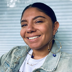 Sylvia Hadnot smiles warmly. She has light brown skin and her black hair is parted and pulled back. She wears hoop earrings, denim jacket, white shirt.