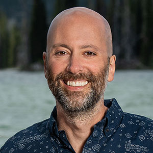 Jason Kanov has a bright smile, brown eyes, white skin, dark beard and a bald head. He wears a blue patterned shirt.