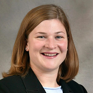 Mary Sass is smiling and has white skin, shoulder-length auburn hair, and wears a black suit jacket and white blouse.