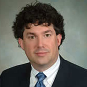 Christopher Kirkey has white skin, brown eyes, and curly black hair. He wears a suit and tie.