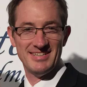 Jason Morris smiles warmly. He has white skin, short brown hair, and wears glasses and a black blazer and white shirt.