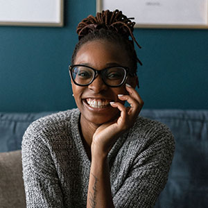 Ani Inyo is smiling enthusiastically. She has black skin and braided hair. She wears large black-framed glasses and a grey knit sweater.