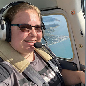 Elizabeth Gibbs is flaying an aircraft and seated in the cockpit. She is smiling, has white skin, and wears a headset, sunglasses and  T-shirt.