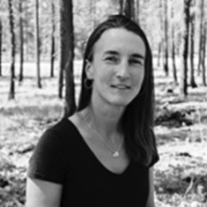 In this black and white portrait, Susan Prichard stands with forest trees behind her. She has white skin, long dark hair, and wears a pendant necklace and black t-shirt.