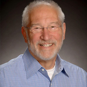 Paul Merriman smiles warmly. He has white skin, grey hair and beard, and wears glasses and a blue shirt.