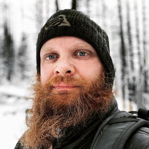 Jonathan Batchelor wears a black ski cap, winter coat, and backpack. They have white skin and a long reddish beard.