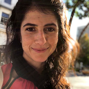 Amani Hariri has a warm smile, large brown eyes, long brown hair, and white skin. She is wearing a pink blouse and black neck scarf.