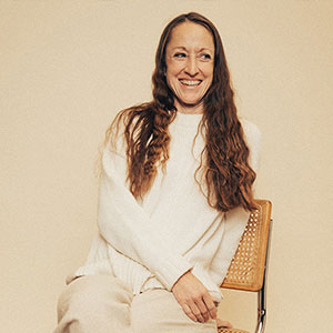 Paula Airth is smiling and is seated on a wicker chair. She has long, wavy, dark hair, white skin, and wears a cream colored sweater.