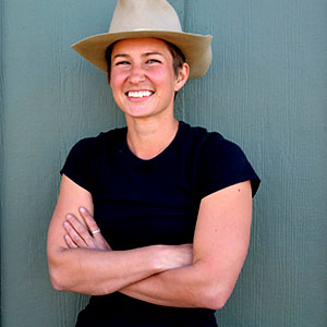 Elibabeth Hayes smiles warmly. She has white skin, her arms are folded, and she wears a black T-shirt and Stetson hat.