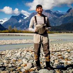 Chris Sargeant is a bearded white male. He stands in a rocky river bed with mountains behind him. He wears rubber boots and overalls.