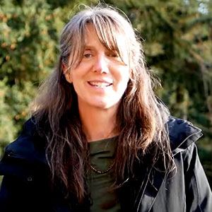 Misty MacDuffee is a white woman. She has long grey brown hair with bangs. She smiles warmly and is wearing a green shirt and black rain jacket.