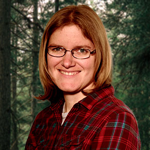 Monika Wieland Shields is a white woman with shoulder length brown hair. She smiles warmly and is wearing glasses a a red plaid shirt.