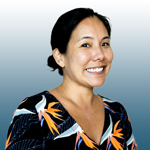 Dr. Kelsie Okamura is a Hawaiian woman. She smiles warmly, has black hair pulled back in a bun, and wears a dress patterned with birds of paradise.