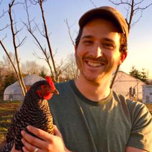 Alex Harris holds a chicken and smiles warmly. Alex has white skin, short facial hair, and wears a T-shirt and baseball cap.