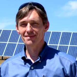 Darrin Magee is a white male with brown hair. He is smiling and wears a blue button up shirt. A large solar panel array is behind him.