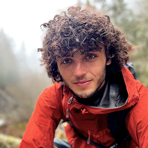 Wil Henkel is a white mail with very curly brown hair and light beard. He smiles and is wearing a red outdoor jacket.