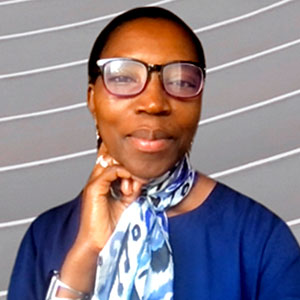 Jeanine Ntihirageza is a black woman with short black hair. She smiles warmly and wears glasses, a dark blue dress, and patterned blue neck scarf.