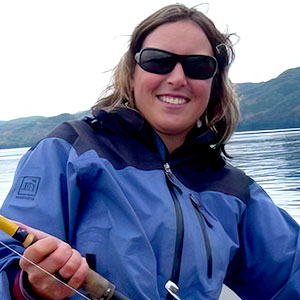 Lindsie Fratus-Thomas is a white woman with shoulder length brown hair. She smiles and wears sunglasses and a blue outdoor jacket. She is holding a fishing rod.