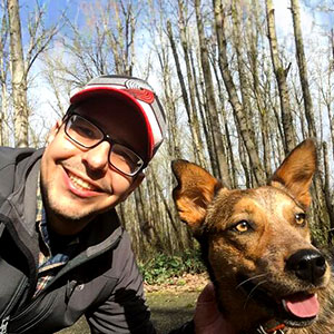 Jaimes Valdez is a light-skinned male. He has a broad smile, wears glasses and a baseball cap and is leaning down to a shepherd dog.