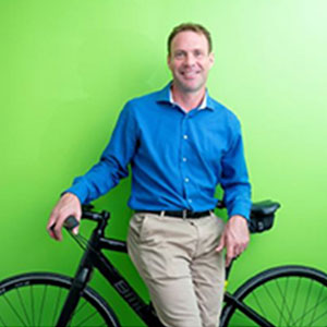 Brian McKenna is a light-skinned male with a warm smile and short brown hair. He wears a blue shirt, chinos, and is standing with his bicycle.