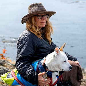 Deborah Giles is a light-skinned female with long sandy brown hair. She wears glasses and a broad-brimmed hat and holds a white dog at her side.