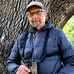 Greg Green is a light-skinned male with glasses, short grey hair and beard. He wears a cap and blue parka and is holding binoculars.