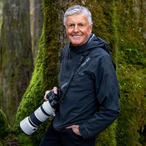 Art Wolfe is a light-skinned male with grey hair. He smiles and is holding a large camera with telephoto lens.