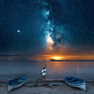 Person on the shore of a beach between two canoes at night beneath a beautiful milky way galaxy
