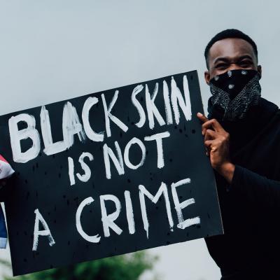 Picture of a Black man wearing a black bandana and long, sleeved shirt holding a sign that reads "Black skin is not a crime". He is also holding an American flag in one hand. In the upper right corner is a WWU blue ribbon with the Alumni Association logo.