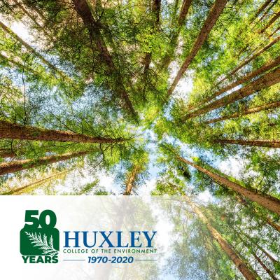 View from base of old growth trees looking up to a bright sky and Huxley 50th logo on the lower left