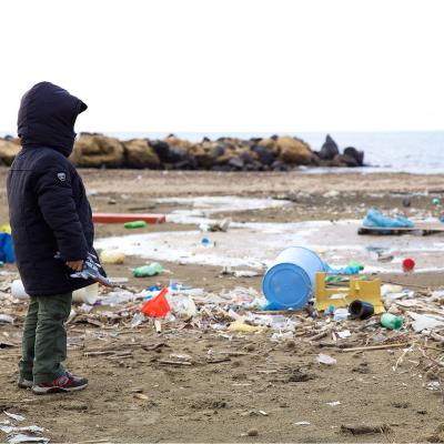 A child standing on a beach looking at trash scattered all over the sand