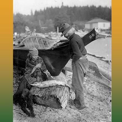 Coast Salish woman seated with a dog and standing man next to traditional fishing vessels
