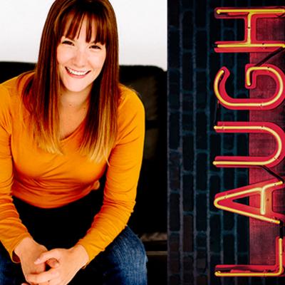 Comedian Anndrea Entz has a bright smile, long chestnut hair with blonde highlights and bangs. Her shirt is bright orange. Next to her is a neon LAUGH sign.