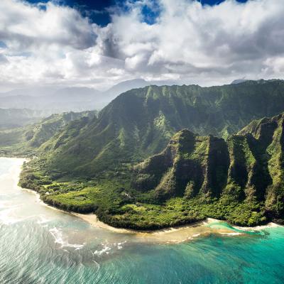 Stunning aerial view of a Hawaiian island with lush green mountain peaks, emerald  ocean water, and deep blue sky with white clouds.